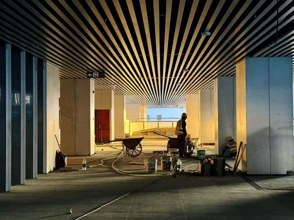 Interior view of a spacious lobby under construction featuring a linear ceiling grid, visible installations and construction workers in the foreground.