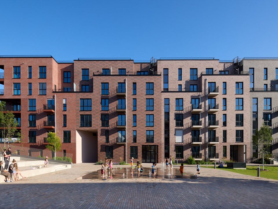 Frontal view of a residential building featuring a rhythmic brick façade, balcony zones and an active district square with a water feature.
