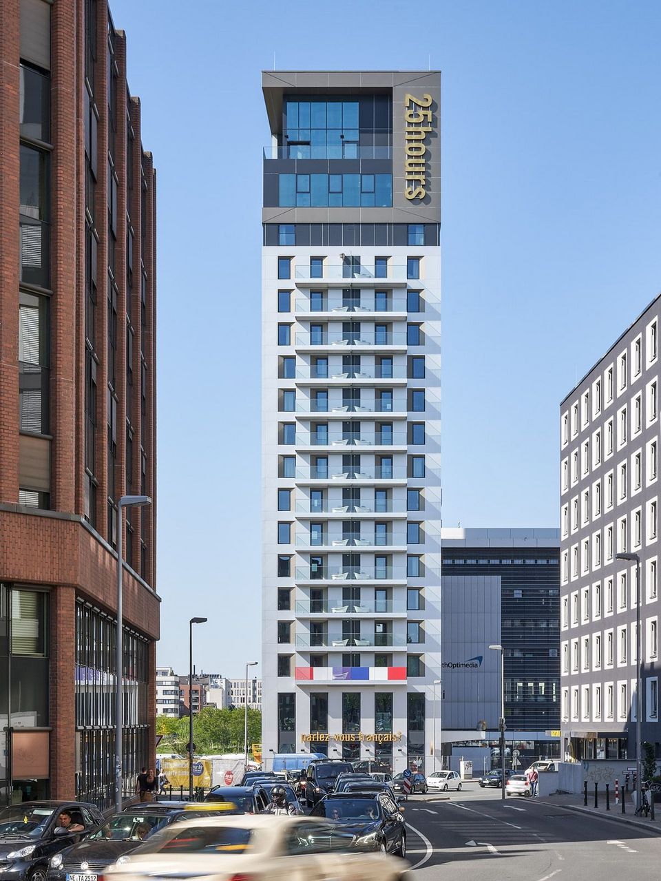 Frontal view of a slender high‑rise building in an urban street setting featuring a structured façade, a cantilevered roof edge and active traffic in the foreground.