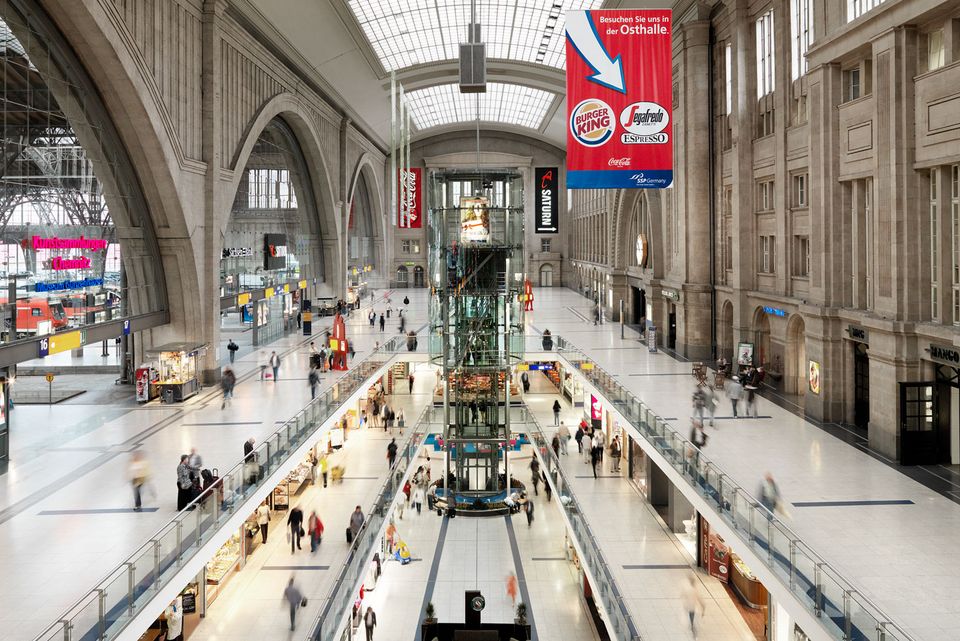 Central perspective view of a main railway station with multi-storey retail areas, glass lift shaft, spacious hall structure and functionally organised routes for passenger and visitor flows.